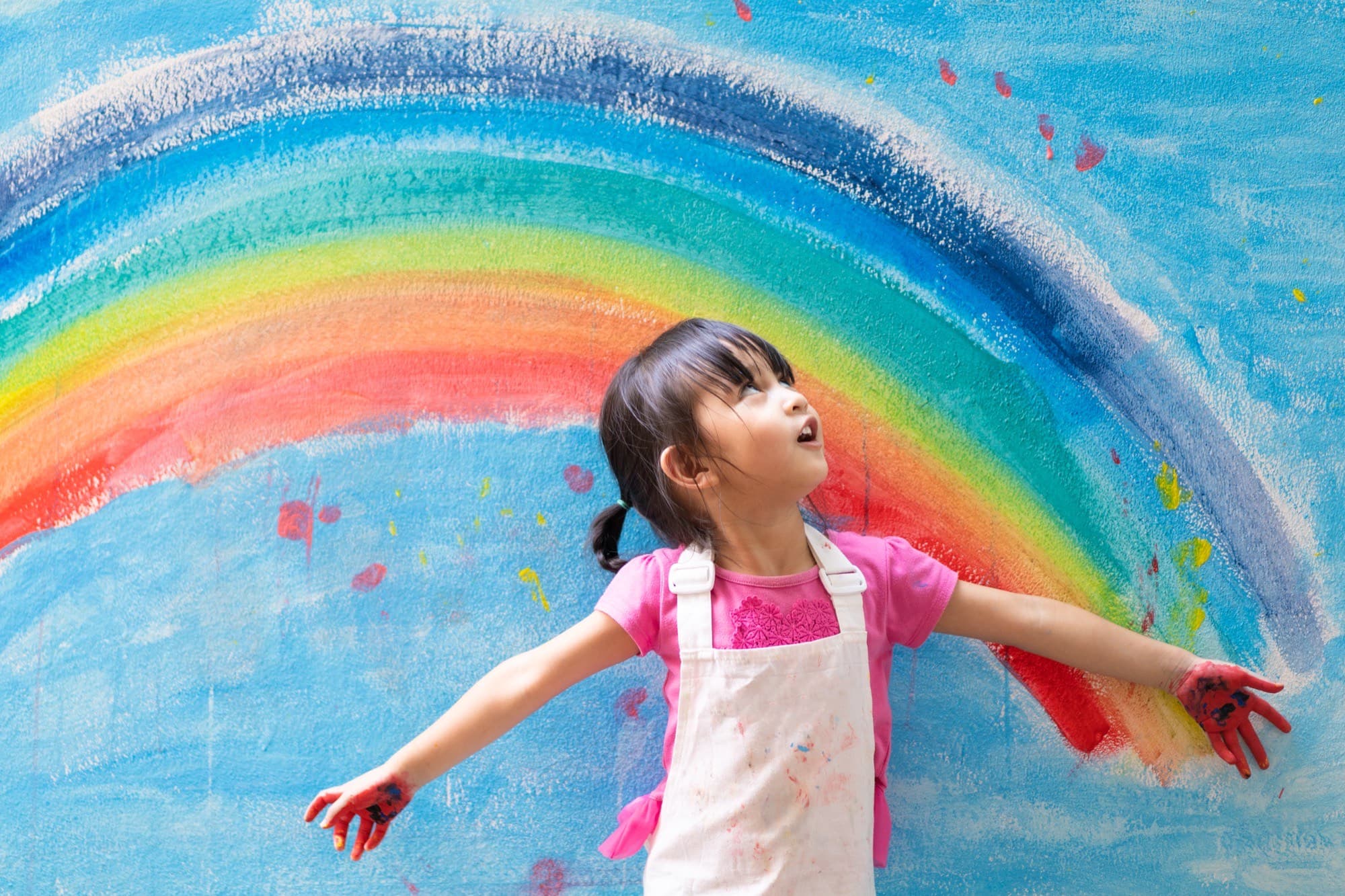 A young child with paint on their hands stands with arms outstretched in front of a large, brightly painted rainbow.
