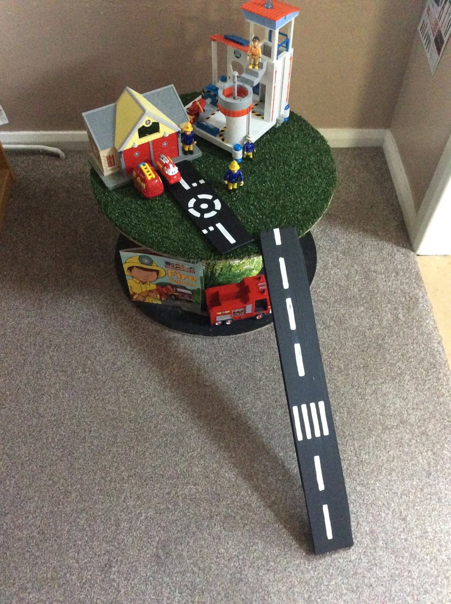 A small-world play set with a toy fire station and road laid out on a grass-topped table.