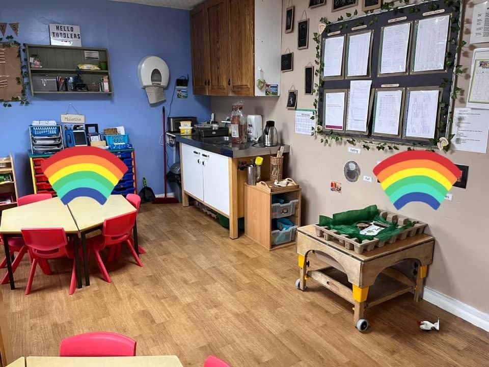 A bright room with painted rainbows on the wall, red tables and chairs and a wooden mud-kitchen trolley.