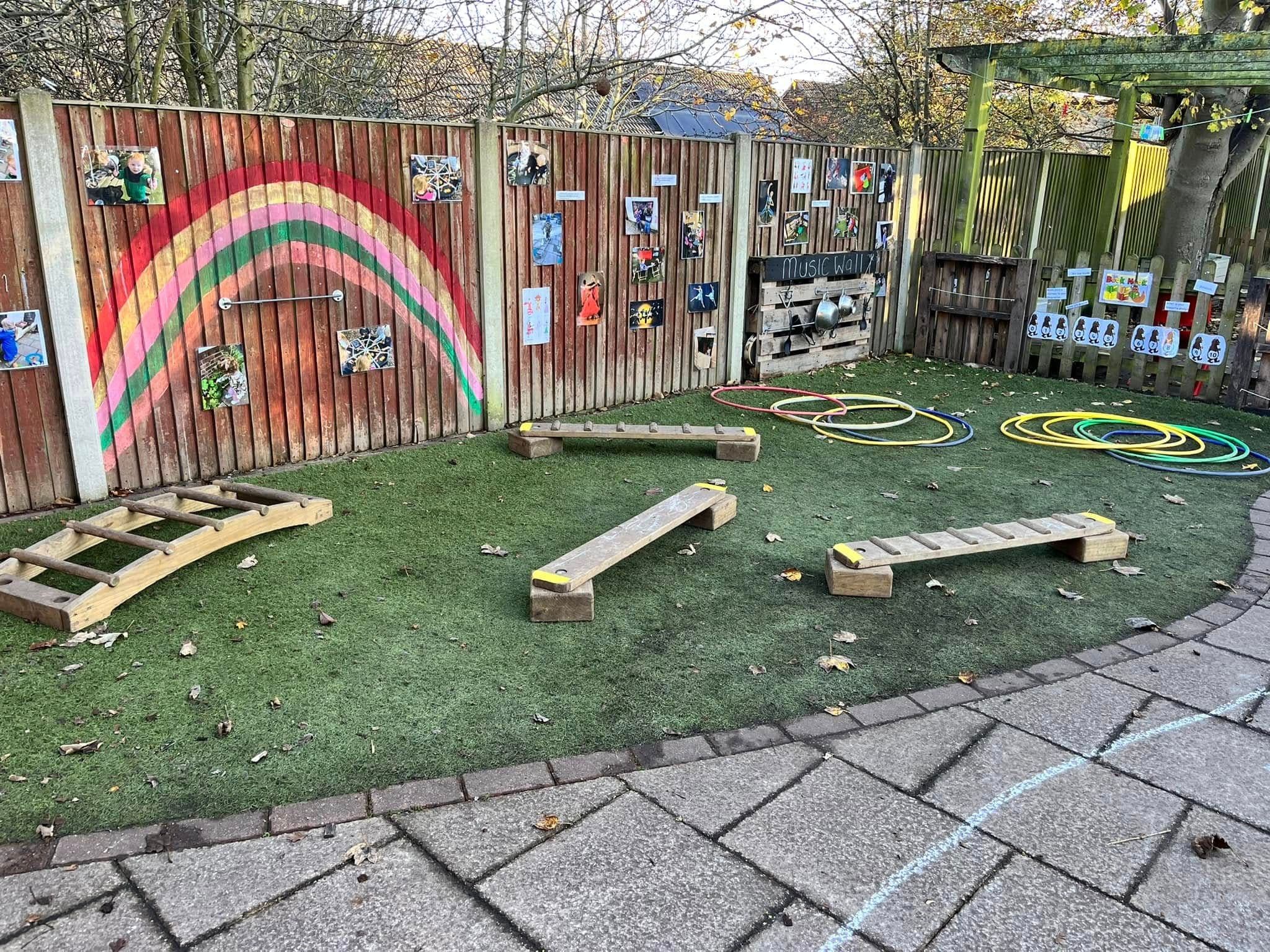 An outdoor play area with a large rainbow painted on the fence and wooden balance beams on artificial grass.