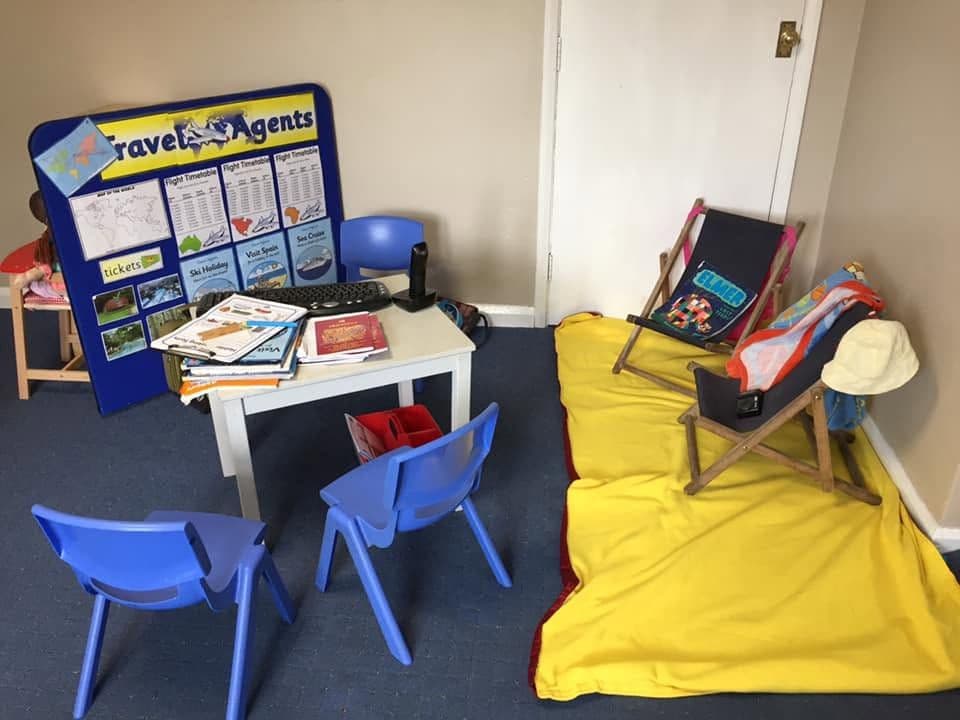 A role-play travel agent's corner with a desk, holiday brochures, a laptop and a striped deckchair.