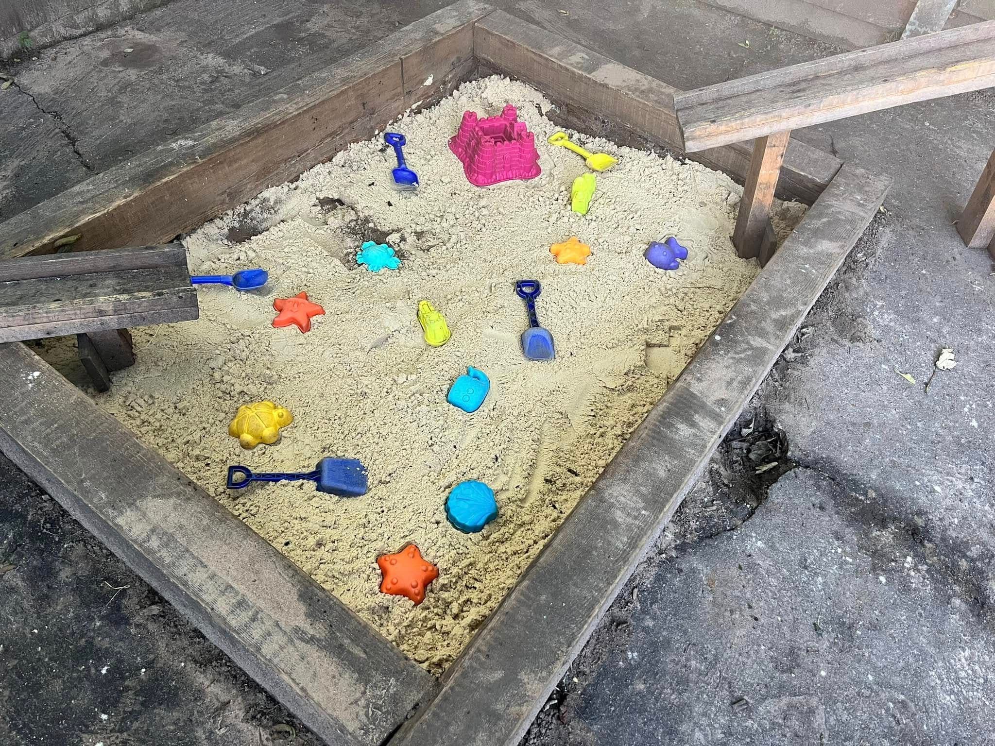 A wooden sandpit filled with sand, moulds and coloured spades.