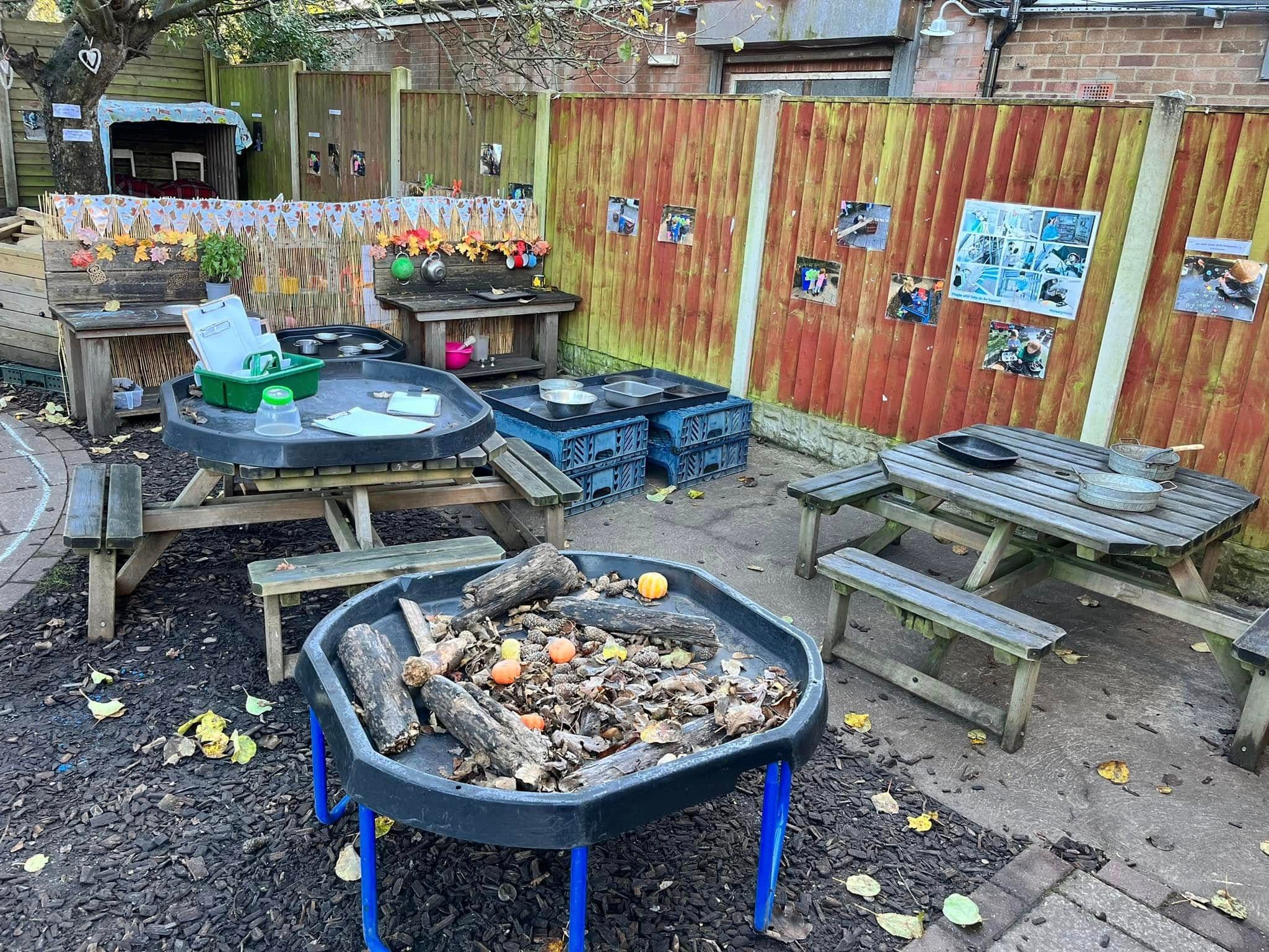 An outdoor learning area with wooden picnic benches and a tuff tray of logs and pumpkins.