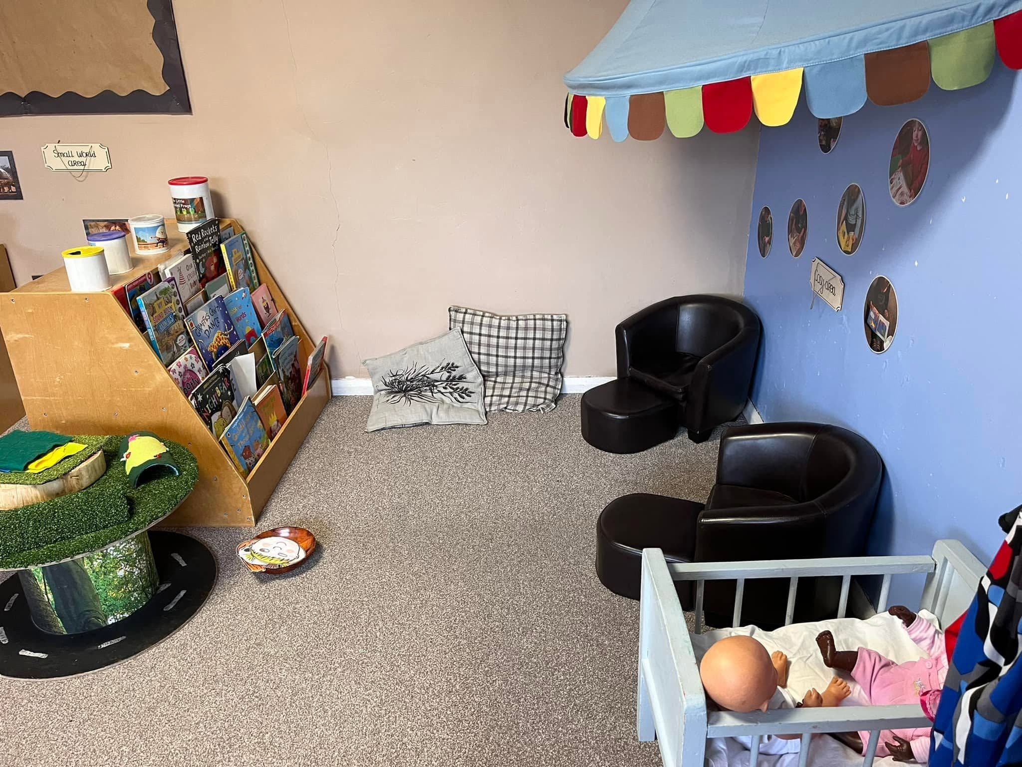A cosy book corner with a fabric canopy, soft tub chairs, a bookshelf and a doll in a cot.
