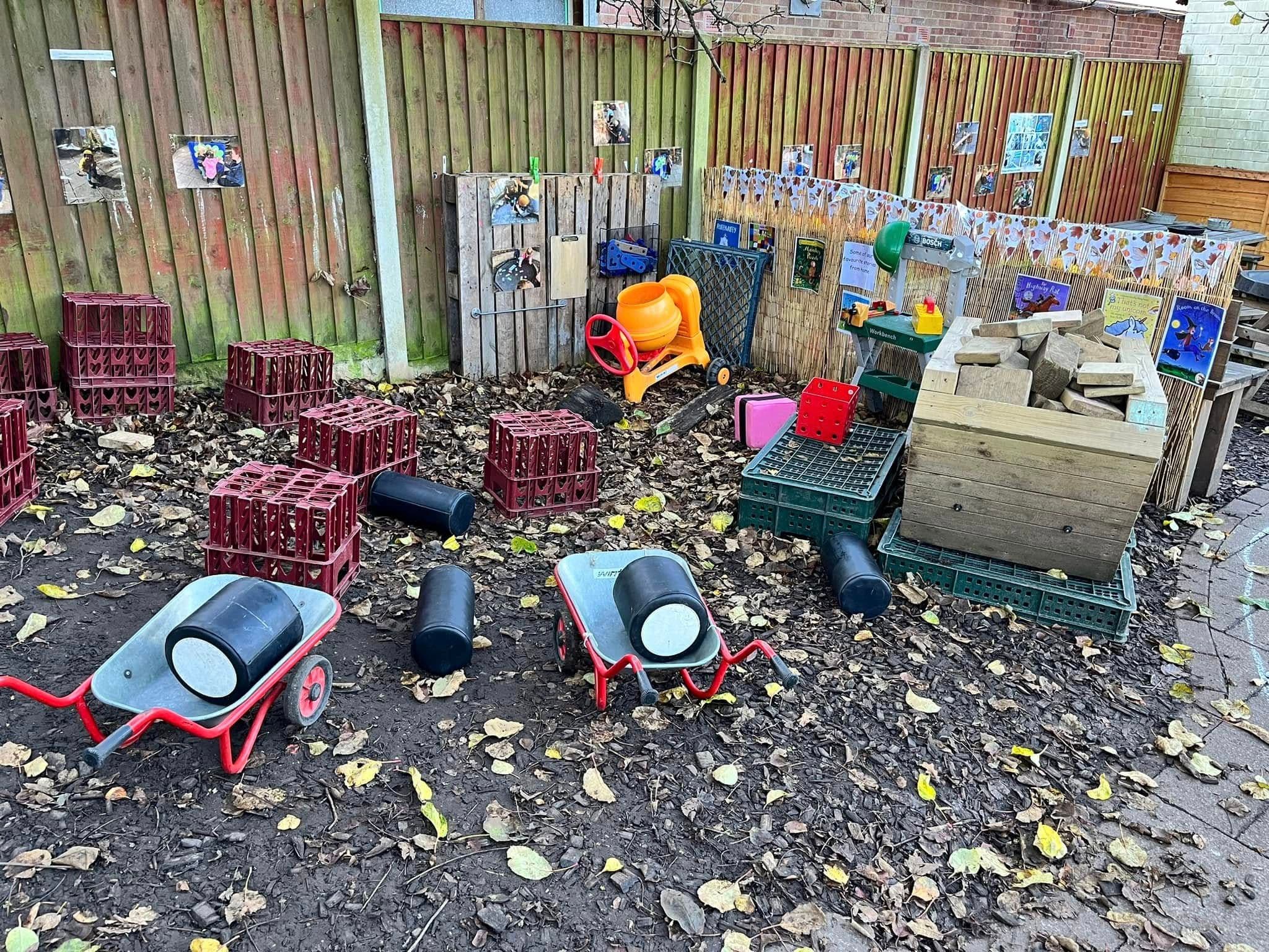 An outdoor mud-play area with toy wheelbarrows, crates and a water wall against a wooden fence.